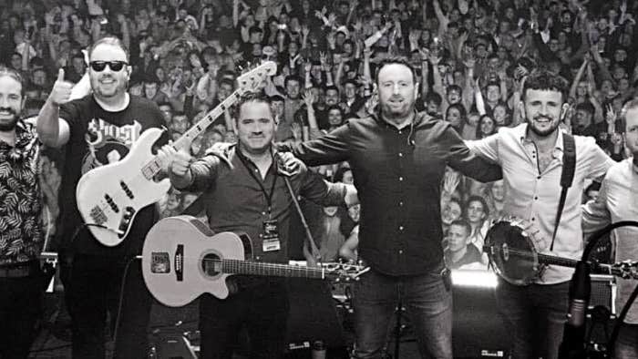 A line of 6 smiling men on a stage with their backs to a large audience.