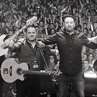 A line of 6 smiling men on a stage with their backs to a large audience.