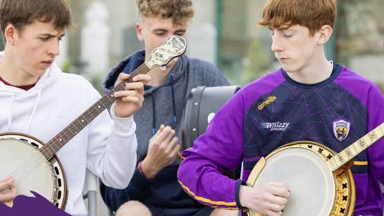 3 young men seated playing musical instruments outdoors, all looking down, concentrating.