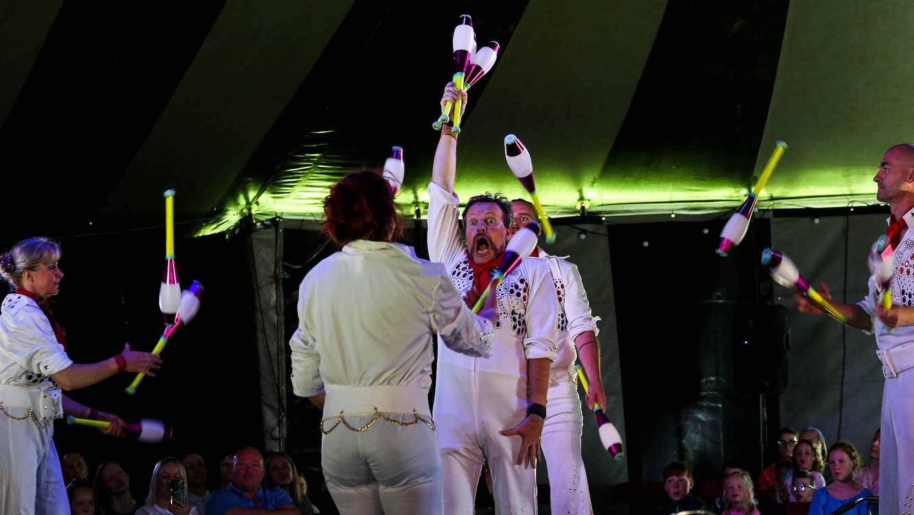 In a dark tent, people dressed in white are juggling in formation with large batons.