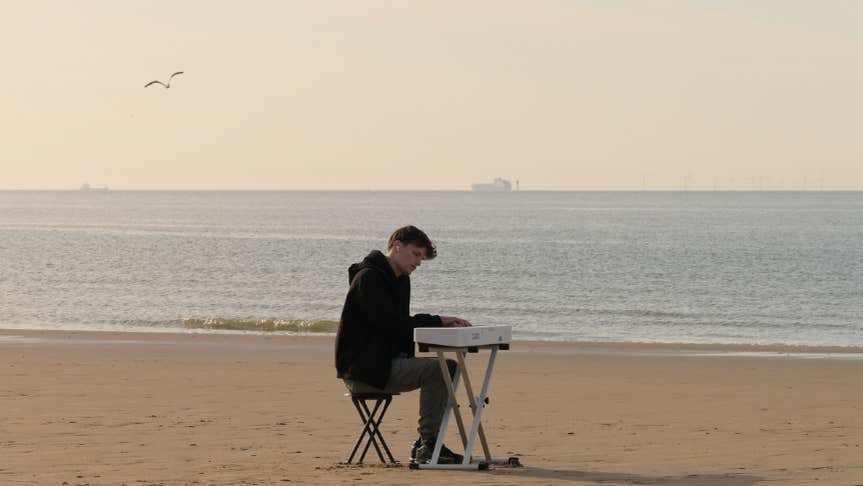 James Malikey - Lost Within The Aether, a man seated at a small portable keyboard on an empty beach