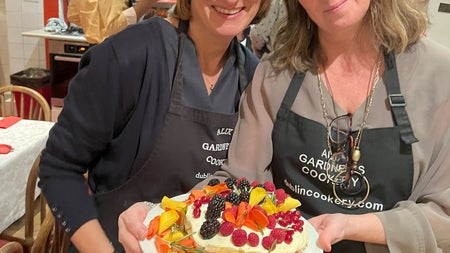 Two ladies in aprons posing with a cake with lots of fruit as decorations