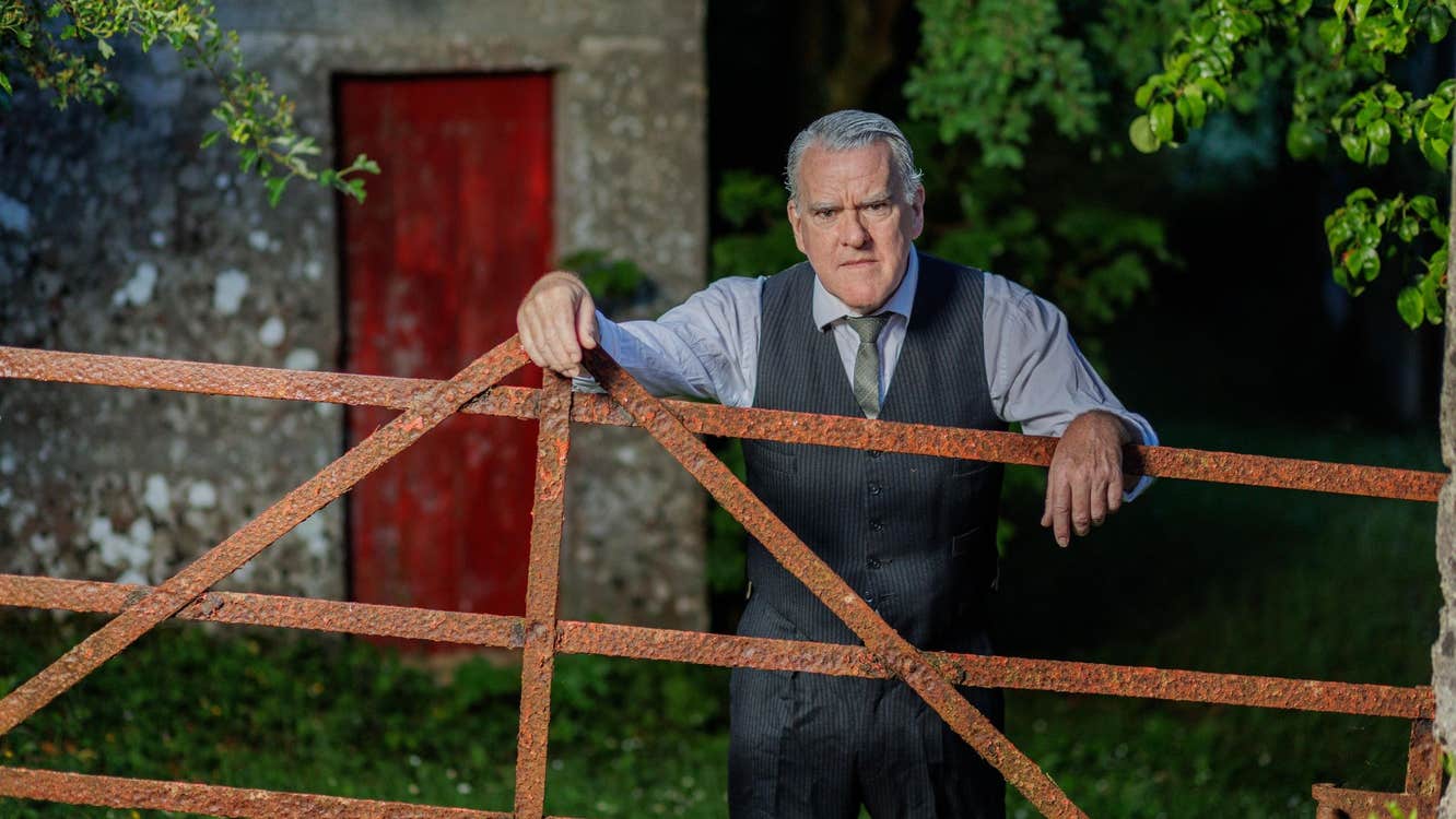 A rather cross looking older man in shirt, tie and waist coat is leaning his arms on a rusty gate