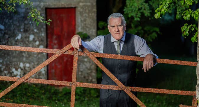 A rather cross looking older man in shirt, tie and waist coat is leaning his arms on a rusty gate