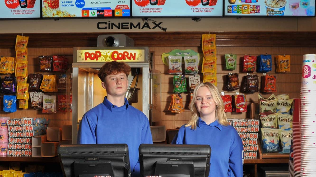 Two people standing behind a wooden counter with packets of sweets a popcorn machine and light up sign advertising cinema snacks