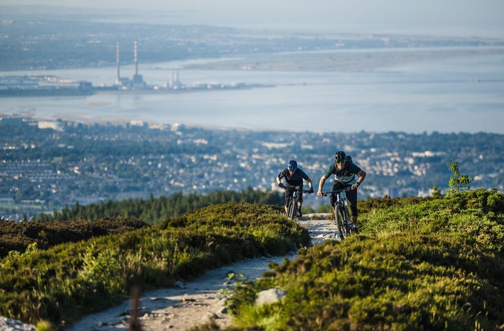 Two cyclists on a biking trail with a view of a city and bay in the background