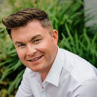 A slightly smiling man in white shirt with grasses in the background.