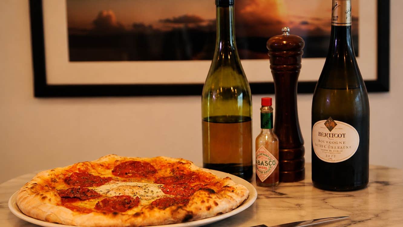 A pizza served alongside a bottle of wine with olive oil and hot sauce