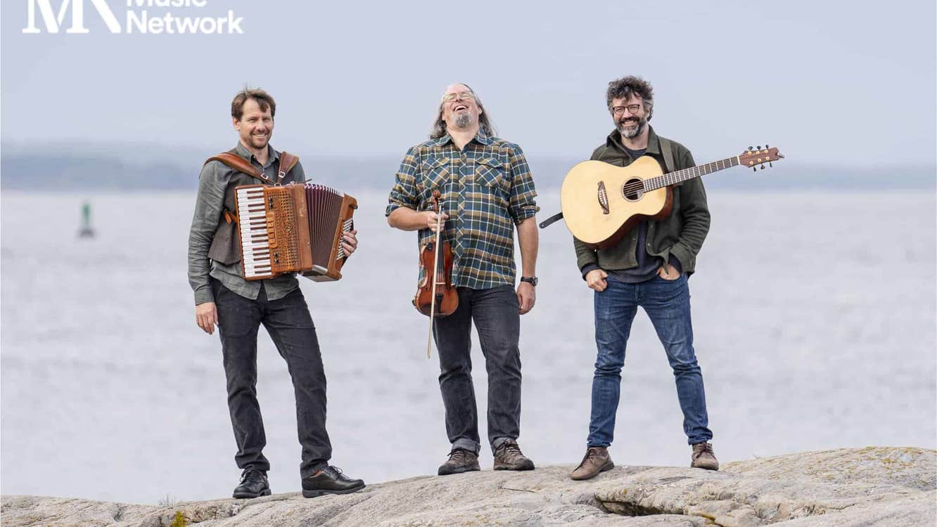 3 smiling men are standing on a large rock with musical instruments against a flat, grey sea.