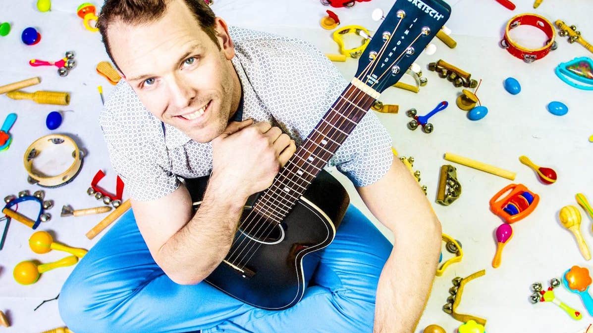 A smiling man holding a black guitar is seated on a white ground surrounded by small, colourful child musical instruments.