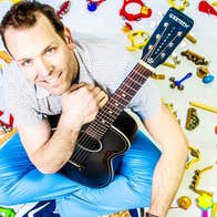 A smiling man holding a black guitar is seated on a white ground surrounded by small, colourful child musical instruments.