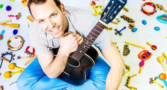 A smiling man holding a black guitar is seated on a white ground surrounded by small, colourful child musical instruments.
