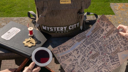 Light brown backpack with a mystery city label on it next to a black notebook with a thread spool and two peoples hands one holding a map and one holding a small box and a magnifying glass