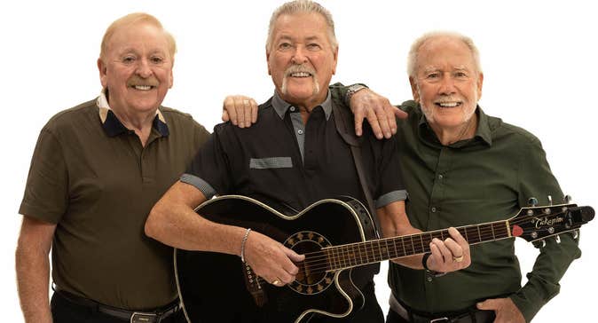 3 older men in a line, the middle one playing a guitar, all smiling, against plain white background.