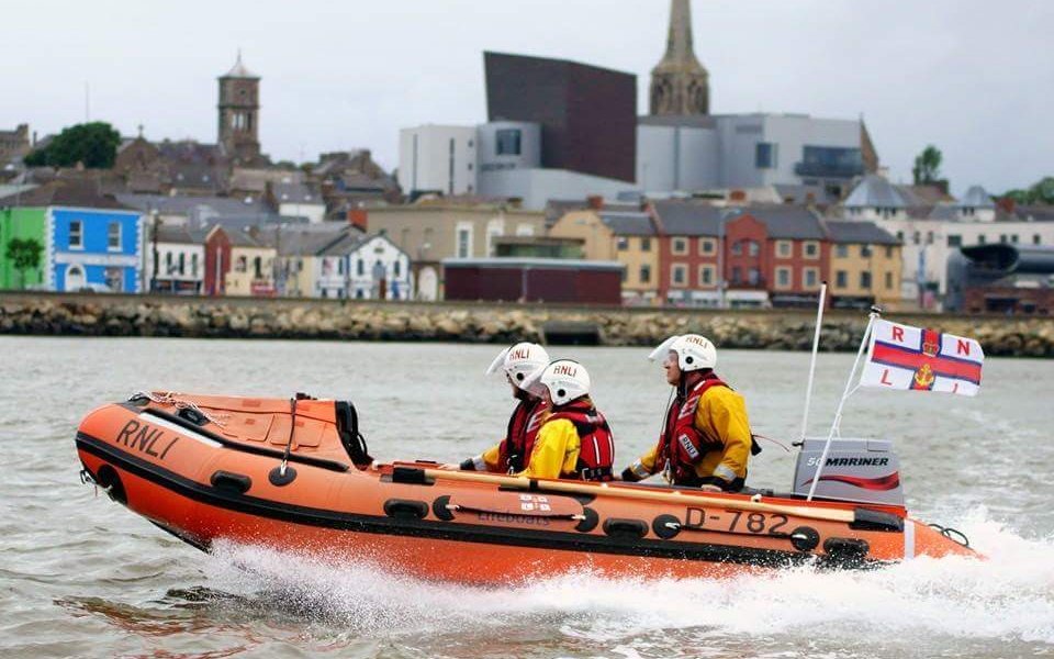 Side view of small, orange inflatable craft with 3 yellow and red lifeboat crew on board looking forward, on grey water with quay and buildings in the background.