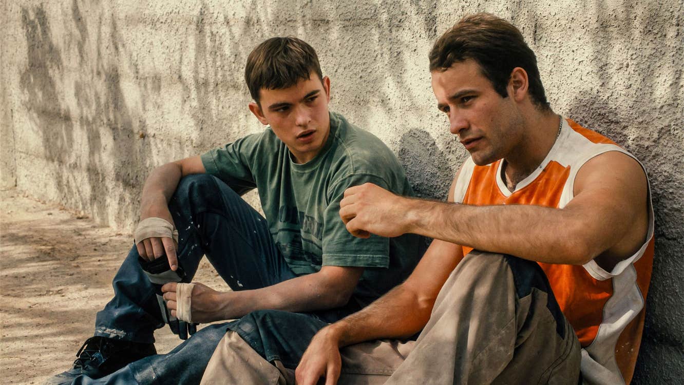 A boy in his late teens and a young man are sitting against a wall.