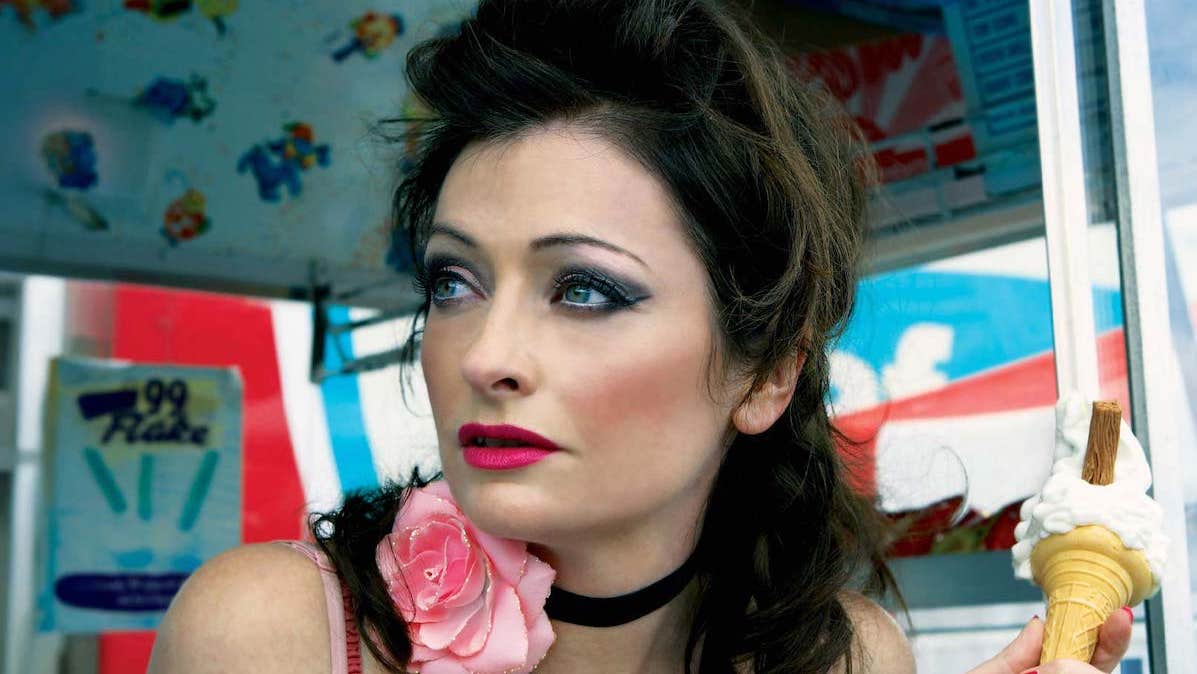 A woman with dark make-up is holding an ice cream looking away to her right.