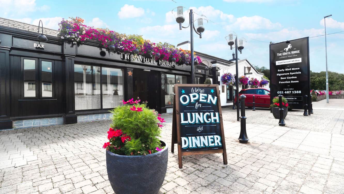 The exterior of a pub with flowers along the front and a lunch sign on the street