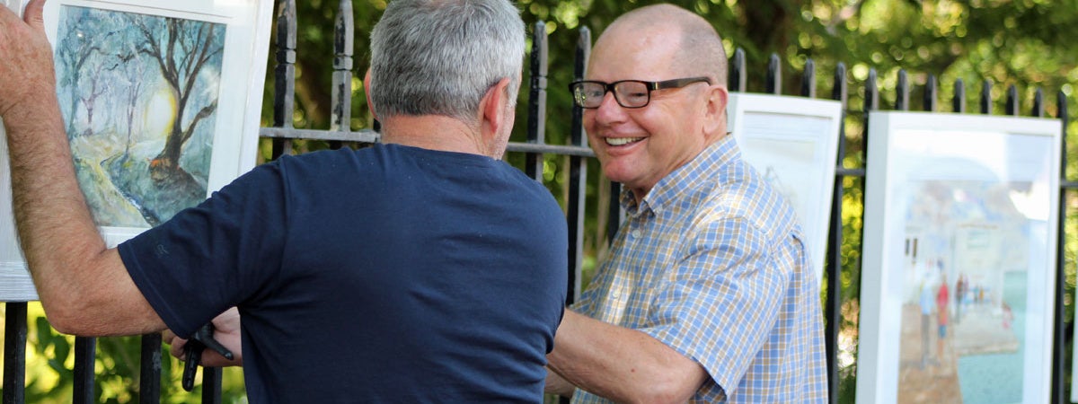 Two men engaged in conversation as one of them hangs a painting on the railing