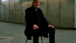 A seated man in dark overcoat is singing with his eyes closed in an empty, concrete covered space.