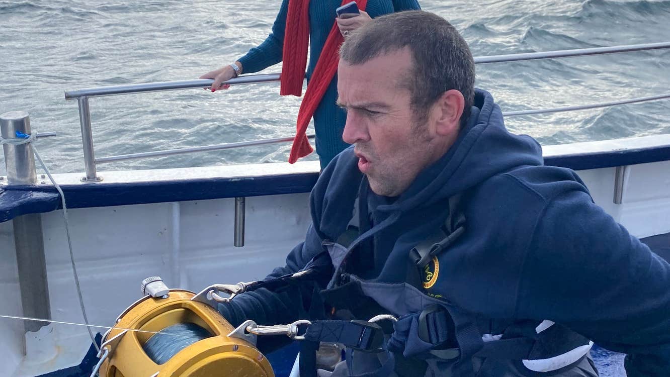 Man on a boat holding a yellow fishing reel with a woman holding a phone in the background