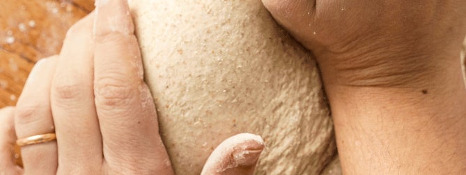 looking down on a pair of hands kneading a large piece of dough on a floured wooden surface.