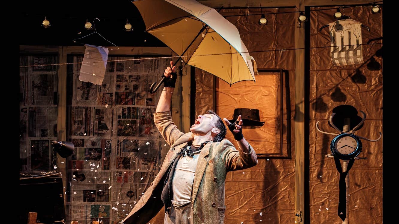 The Collector at An Táin, a man is holding an umbrella over his head on a stage surrounded by busy background stuff.