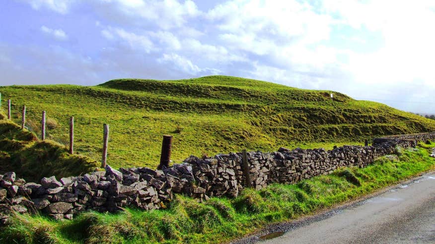 A roadside image of Rathbeg Mound and a stone wall in Rathcroghan