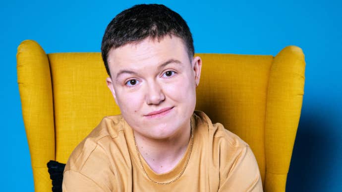Comedian Sinead Walsh, gently smiling, seated in a yellow, highback armchair.