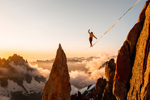 BANFF MOUNTAIN FILM FESTIVAL WORLD TOUR. Photo of high up in red tinged rocky mountain tops, a person is walking along a tightrope between two points with clouds below.