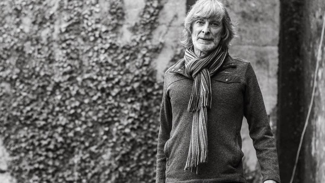 Black and white photo of man wearing a scarf and jumper, standing in front of stone building covered in ivy.