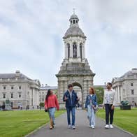 3 people walking on a path with a tour guide talking, impressive grand, stone buildings and tour in the background.