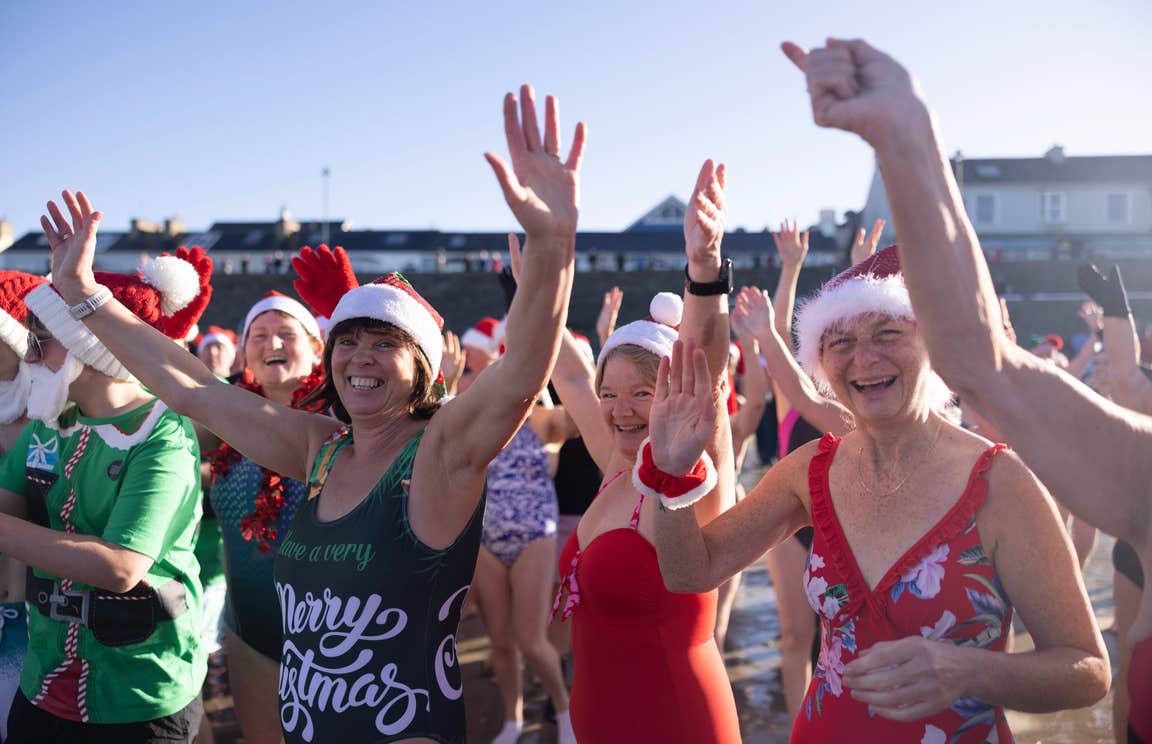 Women in Christmas swimsuits and Santa hats at Kilkee Beach smiling for the camera as they head into the ocean.