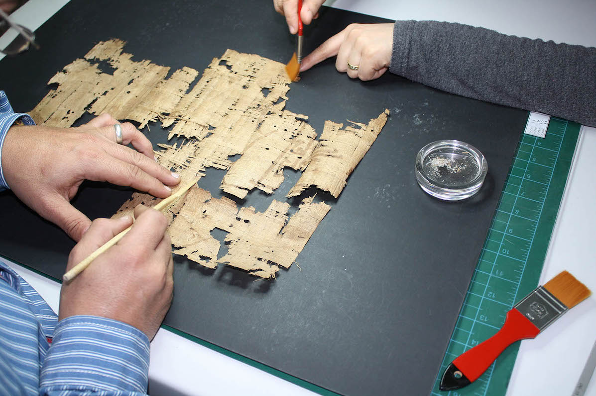 Photo looking down on a board on a table with a very ancient and distressed piece of papyri being attended to with 2 sets of arms visible working with small hand tools.
