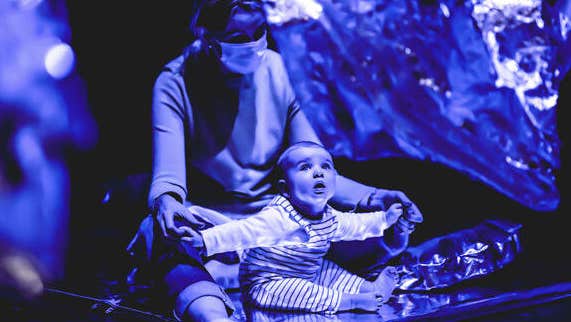 A baby is seated on lap of adult, looking up engaged.