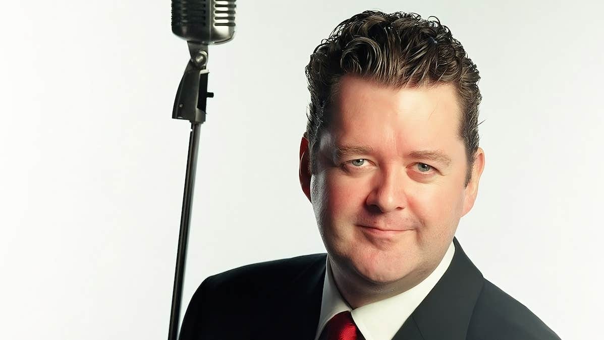 A man in suit and red tie is smiling at the camera, old style large mic is beside him.