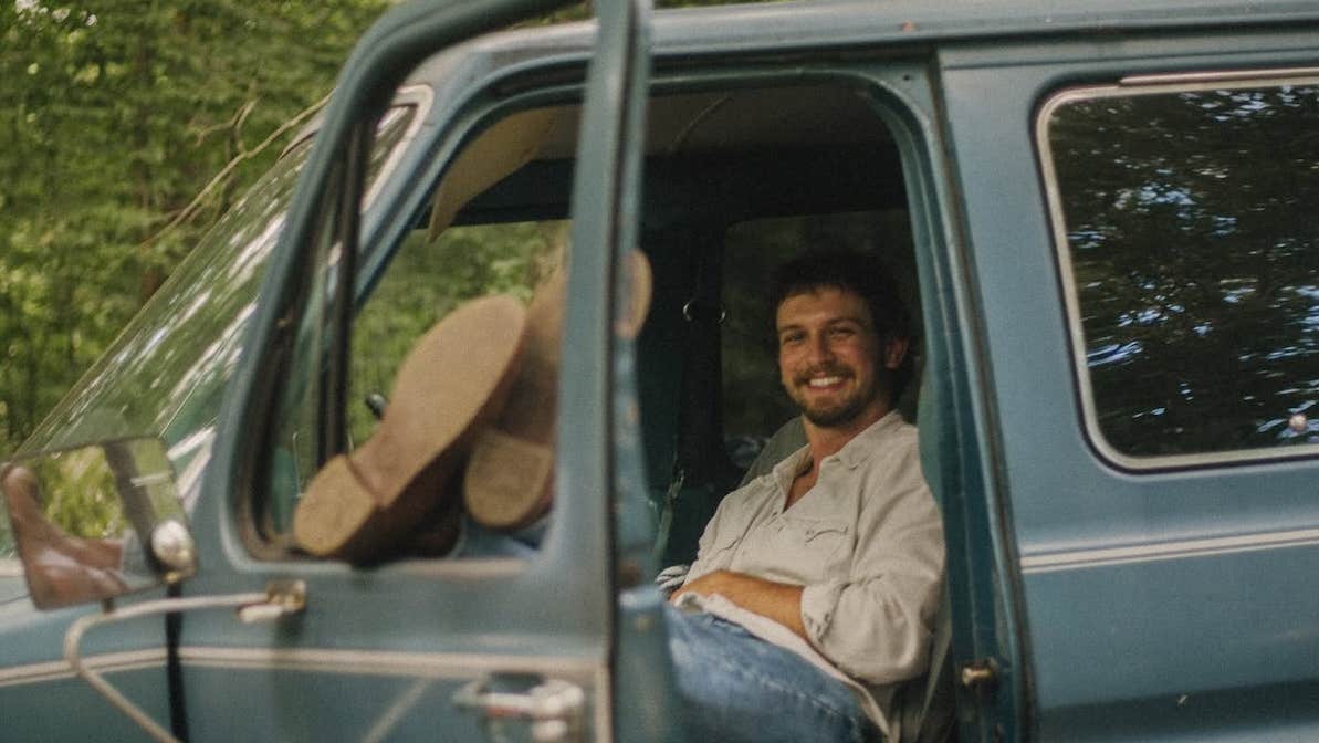 A smiling mans seated in an old van with his legs resting up on the open window of an open door.