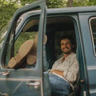 A smiling mans seated in an old van with his legs resting up on the open window of an open door.