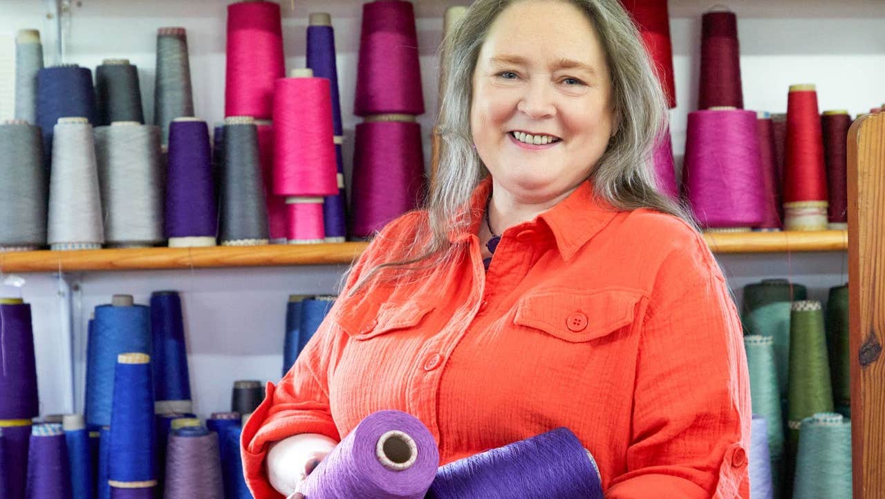 A weaver standing in front of bobbins of different coloured yarns
