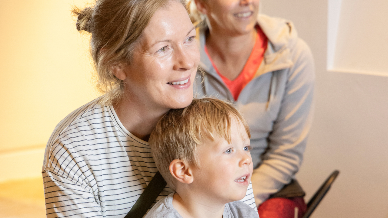 A child and adult are looking at something with interest with another adult in the background.