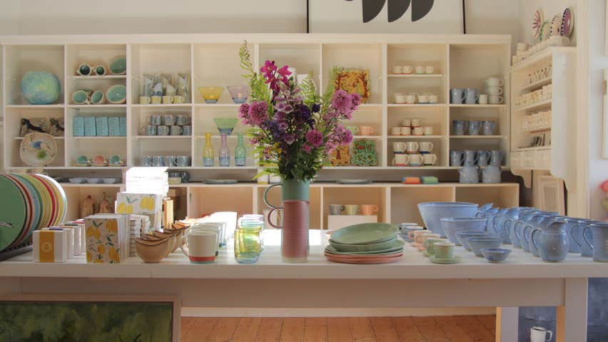 A table with an assortment of pottery with shelves in the background also full of pottery