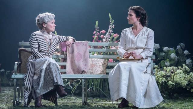 2 women in long dresses are seated on a bench in conversation on a stage