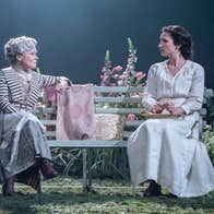 2 women in long dresses are seated on a bench in conversation on a stage