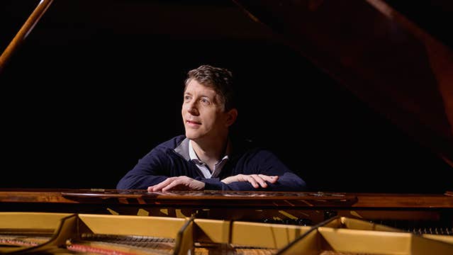 Looking across an open grand piano with a man sat at the keyboard end, looking away, resting his hands on top