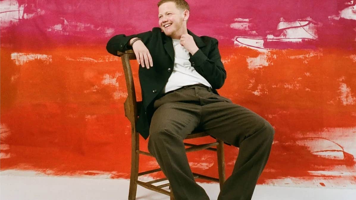 A laughing person in dark jacket and trousers is leaning back on wooden chair against roughly painted red background.