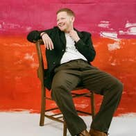 A laughing person in dark jacket and trousers is leaning back on wooden chair against roughly painted red background.
