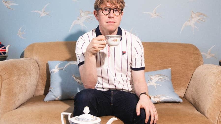 A serious looking man seated on a small sofa holding up a cup of tea with a teapot in front on a low table