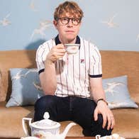 A serious looking man seated on a small sofa holding up a cup of tea with a teapot in front on a low table