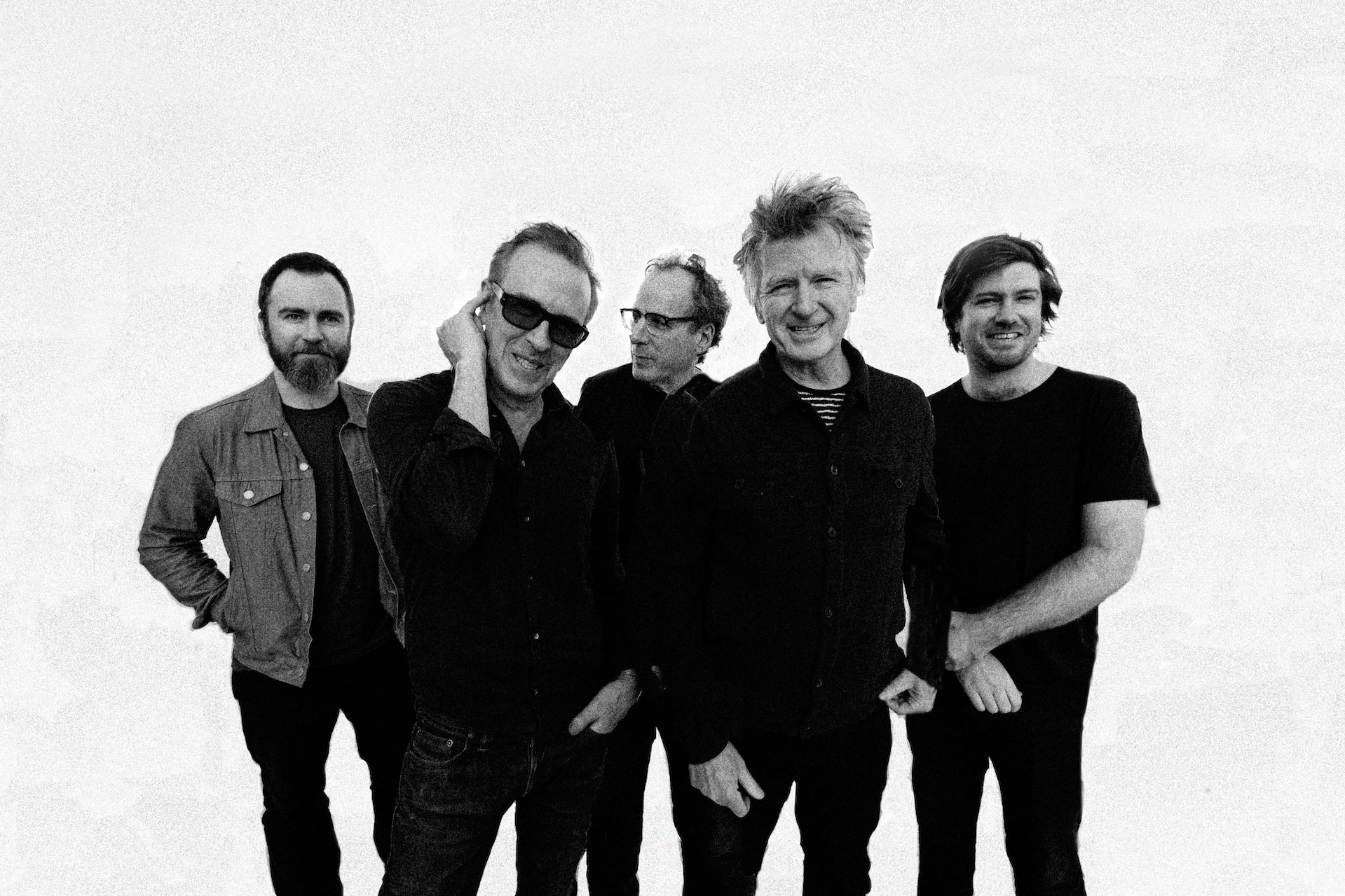 Black and white photo of 5 men dressed in black standing in a loose group, mostly smiling, against plain white background.