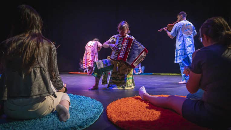 Musicians moving around a stage whilst playing to a seated adult and baby
